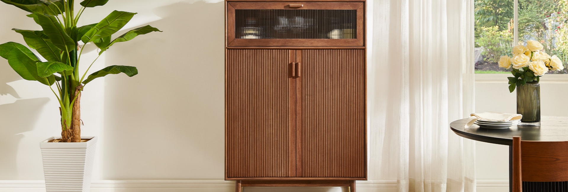 A MCM bar cabinet stands centered between a plant on the left and a dining table with a vase of roses on the right.