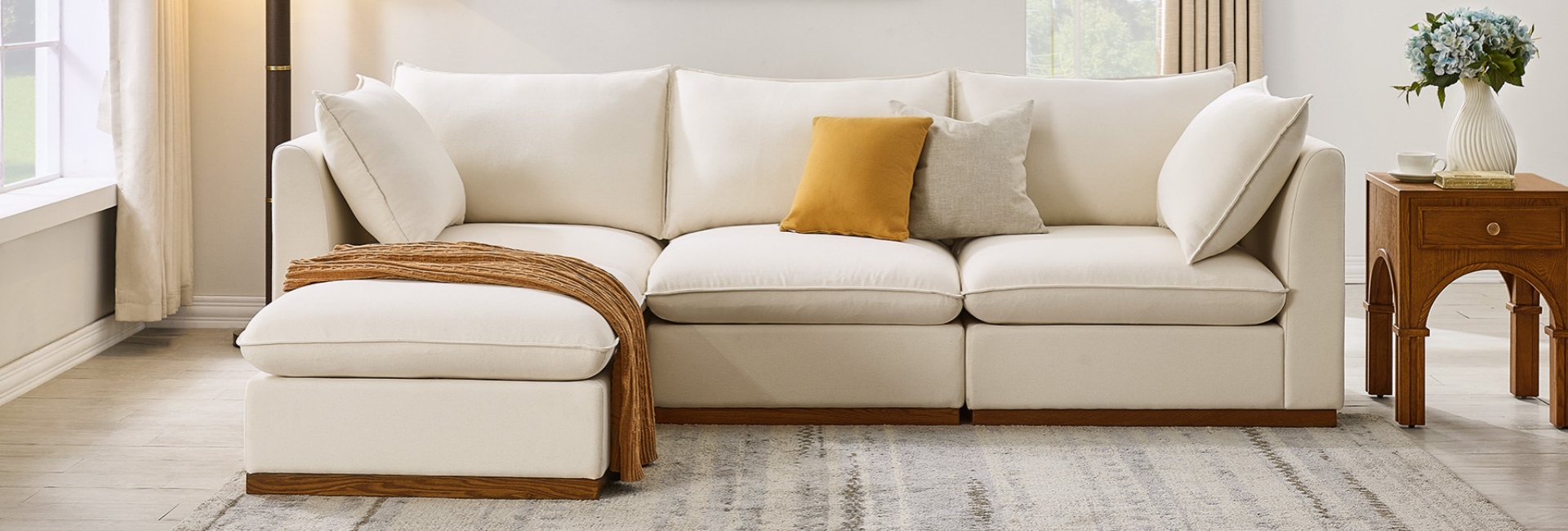 A modern cream modular sectional sofa with an ottoman, featuring a solid wood plinth base and soft coastal-style pillows.