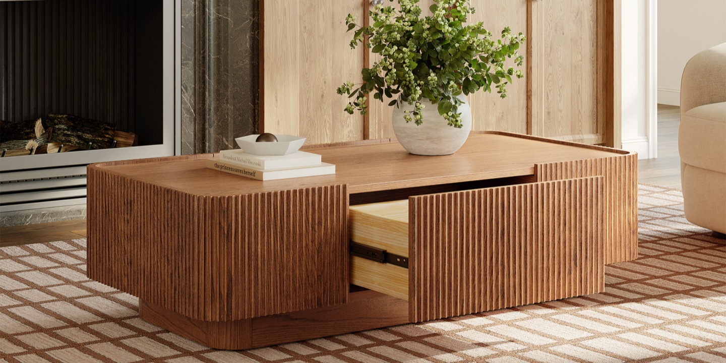 Skimra 55 inch Oak Coffee Table featuring distinctive reeded (fluted) wood sides with soft rounded corners. The surface displays a white geometric bowl, books, and a large white vase with green foliage.