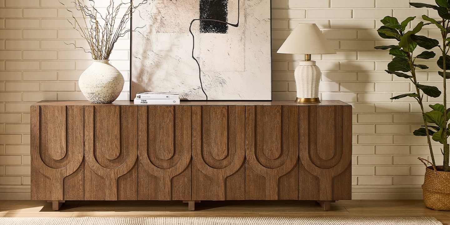 An Hernest 84 inch oak media console with patterns styled with a table lamp, ceramic vase, artwork, and a potted plant. An Hernest 84 inch oak media console with patterns styled with a table lamp, ceramic vase, artwork, and a potted plant.