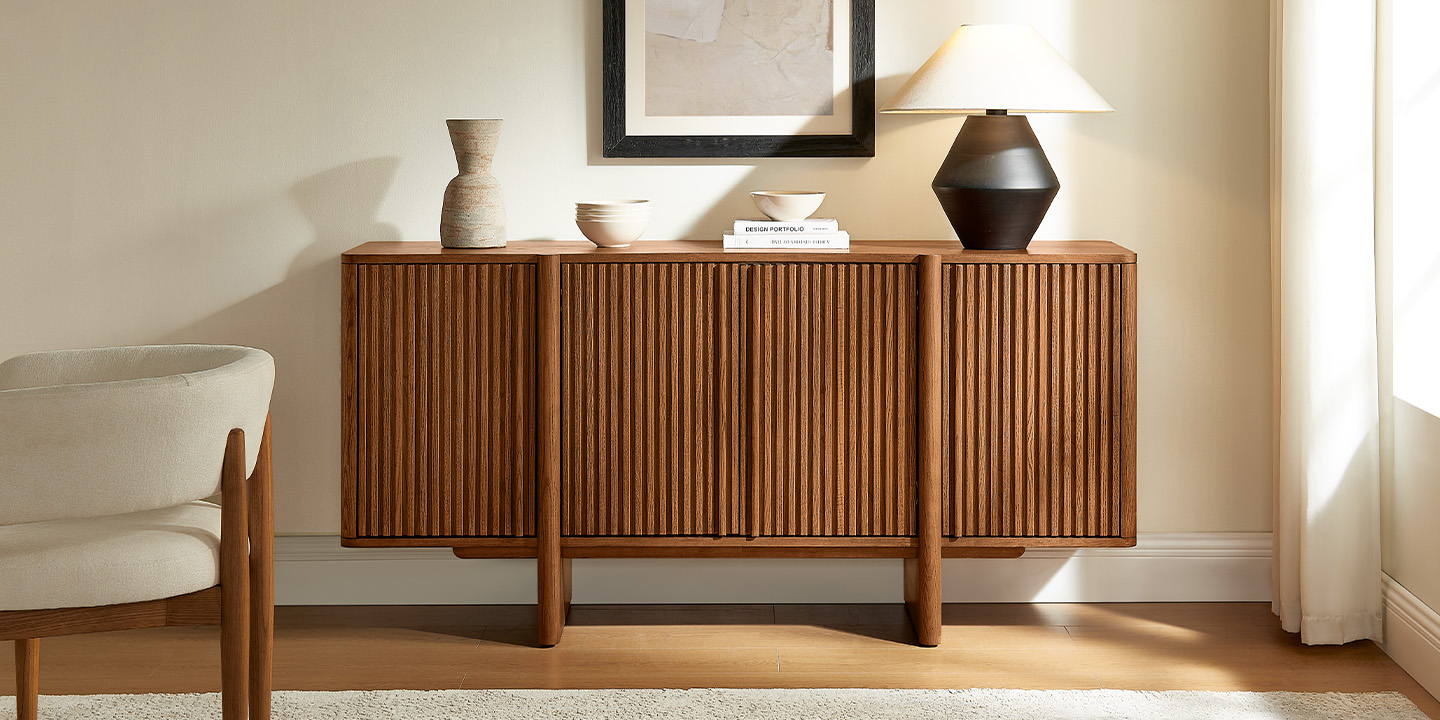 An ash wood fluted sideboard topped with a ceramic vase, books, and a lamp, styled next to a modern armchair. An ash wood fluted sideboard topped with a ceramic vase, books, and a lamp, styled next to a modern armchair.