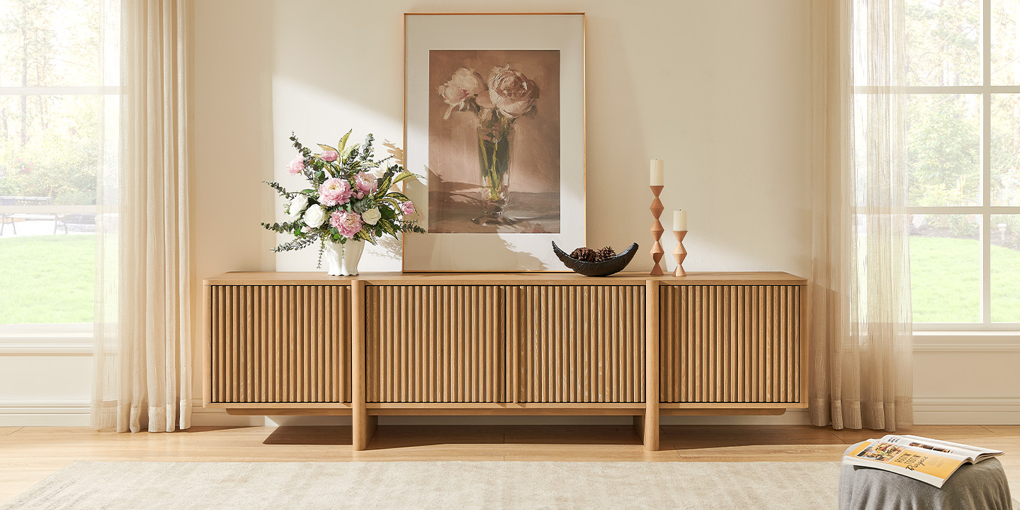 Natural oak fluted media console styled with vibrant pink flowers, a framed floral painting, and candle holders.