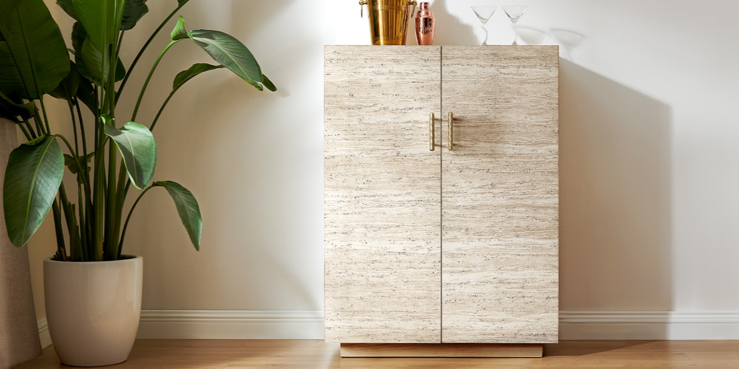 The Renata 36-inch travertine bar cabinet, with brass-toned hammered hardware, stands next to a large potted plant. The Renata 36-inch travertine bar cabinet, with brass-toned hammered hardware, stands next to a large potted plant.