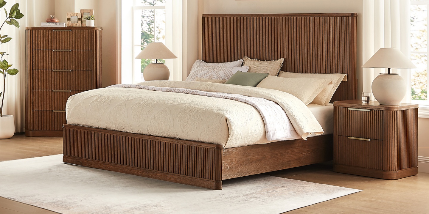 This tall 38-inch Finnley fluted 5-drawer chest features rich brown wood and stands next to a matching bed in a bedroom. This tall 38-inch Finnley fluted 5-drawer chest features rich brown wood and stands next to a matching bed in a bedroom.